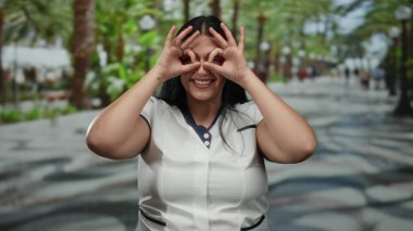 Canlı bir park ortamında neşeli hareketlerle gülümseyen bir kadın, yemyeşil ve palmiye ağaçlarıyla çevrili, güneşli bir günde neşeli bir açık hava sergiliyor..