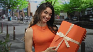 Genç kadın, şehir merkezindeki işlek caddede elinde büyük turuncu bir hediye tutarken gülümsüyor; neşe.