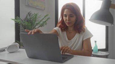 Ev stüdyosundaki beyaz bir masanın üzerine dizüstü bilgisayarı kapatmak için uzanan bir kadın kulaklık ve lamba görünür; uzaktan çalışma konsantrasyonu.