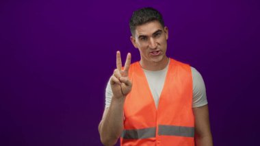 Turuncu güvenlik yeleği giyen mühendis işaret parmağını yukarı kaldırarak mor stüdyoda gülümsüyor..