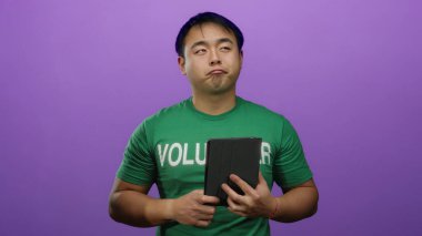 Yeşil gönüllü gömlekli genç adam mor arka plana karşı tablet tutuyor. Toplumun katılımını ve dijital bağlantıyı vurgulayan izole bir ortam sergiliyor..