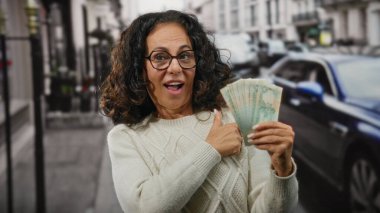 Kadın hareketli bir caddede heyecanlı bir şekilde Dirham banknotlarını tutuyor, neşe dolu bir ifadeyle şehir ortamında pozitiflik ve başarı sergiliyor..