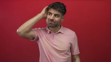 İnsan başını kaşıyarak omzunu parlak kırmızı bir stüdyo duvarına yaslar ve şaşkın bir ifade sunar..
