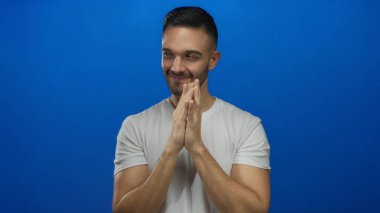 İçerik ifadeli genç bir adam parlak mavi bir arka plana karşı duruyor, elleri sımsıkı bağlanmış, beyaz bir gömlek giyiyor, neşeli ve pozitif bir havayı somutlaştırıyor.