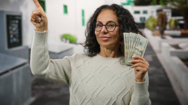 Orta yaşlı kadın Amerikan dolarlarını neşeyle elinde tutuyor şehir sokaklarında başarıyı ve zenginliği işaret ediyor..