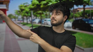 Güneşli bir caddede sakallı genç bir adam yeşil ağaçlarla ve arka planda park edilmiş arabalarla jestlerle bir yeri işaret ediyor..