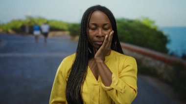 Kıvırcık saçlı, sarı gömlekli, deniz kenarında gezinen bir kadın. Yüzü açık, açık gökyüzünün altında, geçmişi bulanık insanlar.