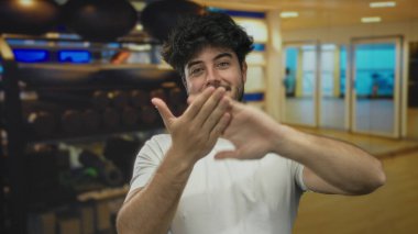 Genç bir İspanyol jesti, spor salonunda para harcayıp, içerideki fitness ekipmanlarıyla çevrelenmiş coşkuyu gösteriyor, finansal jestlerin ve fitness kültürünün kesişimini vurguluyor..