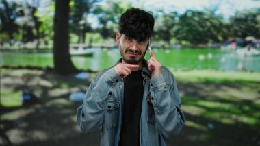 Güneşli bir parkta telefonla konuşan genç bir adam kot pantolon giyiyor, jestlerle düşünceliliğini ifade ediyor..