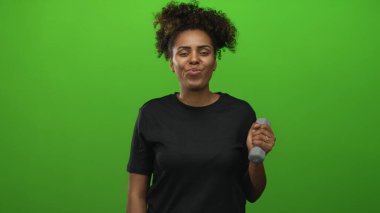 Yeşil stüdyoda dambılı tutarken dudaklarını büzen Afro-Amerikalı genç bir kadın..