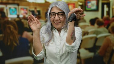 Sanat galerisinde elinde anahtarlarla gülümseyen gri saçlı yaşlı bir kadın. Etrafı insanlarla ve renkli resimlerle çevrili..