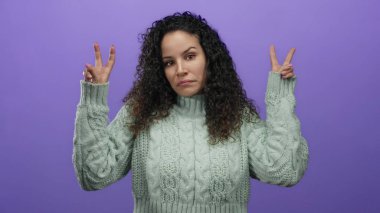 Yeşil kazaklı İspanyol bir kadın parmaklarıyla tırnak işareti yapıyor, mor bir arka plana karşı duruyor, belirsizlik ve iğnelemeyi ifade ediyor..