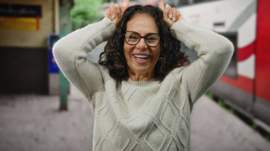 Kıvırcık saçlı bir kadın, dışarıda tren istasyonunda neşeli jestler yapıyor, demiryolu zeminine neşe ve güven saçıyor, gözlük takıyor ve rahat bir süveter giyiyor..