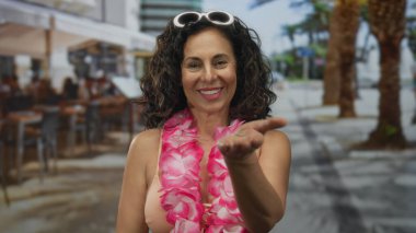 Orta yaşlı kadın restoran terasında kameraya elini uzatıyor; davetlilere sıcak bir karşılama..