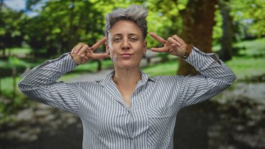 Stüdyoda kısa saçlı ve çizgili gömlekli bir kadın bir gözünü kırparken çift barış işareti hareketi yapıyor..