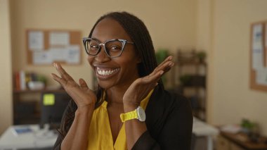 Ofis ortamında gülümseyen gözlüklü mutlu bir kadın. Elleri yüzü havada, neşeli bir ifade sergiliyor. Kapalı bir ortamda, çalışma alanı unsurlarıyla..