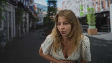 Kadın karnını tutuyor ve öne doğru eğiliyor, kaldırım taşlı bir sokakta dudaklarını büzüştürüyor beyaz benekli bluz giyiyor; karın ağrısı rahatsızlığı.