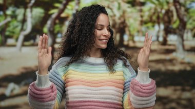 Parkta, açık havada, renkli bir süveter giymiş gözleri kapalı gülümseyen, ağaçlarla çevrili, güneşli bir ortamda el hareketi yapan bir kadın..