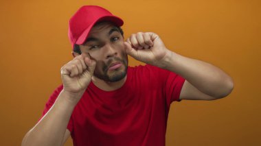 Gözleri ovuşturan ve dudakları çökmüş yanaklara parmak uçlarını bastıran adam turuncu stüdyo ortamında kırmızı şapka ve kırmızı gömlek giyiyor. Üzüntü..