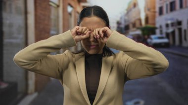 Canlı bir şehir caddesinde gülümseyen ve gözlerini ovuşturan bir kadın, sürpriz ve sevinç taşıyor, bej bir ceket giymiş, canlı bir açık hava ortamında genç coşkusunu somutlaştırıyor..