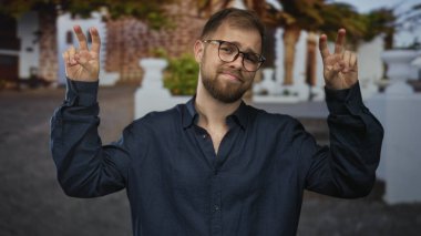 Adam taş bina girişinin önünde iki eliyle barış işareti gösteriyor, gözlük ve günlük gömlek takıyor; oyuncu özgüven ve kolaylık.