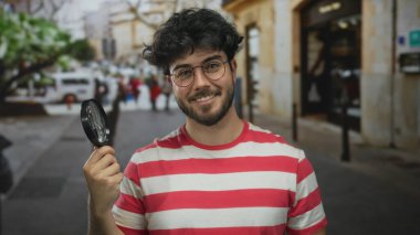 Gözlüklü, sakallı, elinde büyüteç olan kırmızı çizgili gömlek giyen, şehir ortamında gezen Latin kökenli genç bir adam..