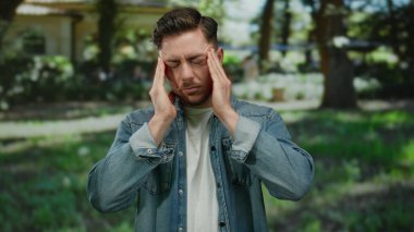 Bir park ortamında başını sakallı genç adam gündüz vakti kot ve beyaz gömlek giyerek açık hava yeşilliğinin ortasında stresli görünüyor..