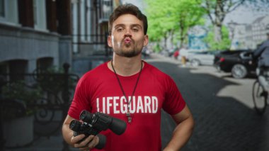 Şehir caddesinde park halindeki arabalar ve ağaçlarla dolu dürbün tutan kırmızı bir cankurtaran tişörtü giyen İspanyol genç adam..