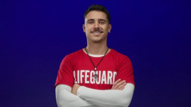 Kırmızı bir cankurtaran tişörtü giymiş İspanyol genç adam izole edilmiş mavi bir arka plan duvarına karşı güvenle kollarını kavuşturuyor, profesyonellik ve hazırlık hissi yayıyor..