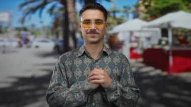 Güneşli bir şehir sokağında arka planda palmiye ağaçları ve bulanık pazar çadırlarıyla kalp hareketi yapan genç bir İspanyol..