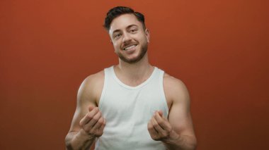 Sakallı genç beyaz adam, izole edilmiş kırmızı arka plan duvarının üzerinden para çekiyor, kolsuz bluz giyiyor, gülümsüyor..
