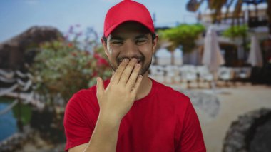Kırmızı gömlekli ve şapkalı adam, sokak restoranının terasında güneş ışığı altında ağzını eliyle kapatıyor..