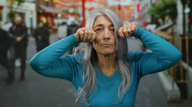 Kıdemli, gri saçlı, mavi kazaklı bir kadın arka planda bulanık yayalarla kalabalık bir sokakta şakacı hareketlerle hayat dolu bir şehir manzarasını yakalıyor..