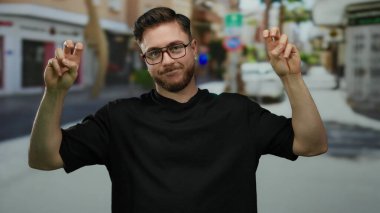 Sakallı genç bir adam, şehir merkezinde gün ışığında dışarıda dikilirken gözlük ve günlük giysiler gösteren bir sokakta barış işareti yapıyor..