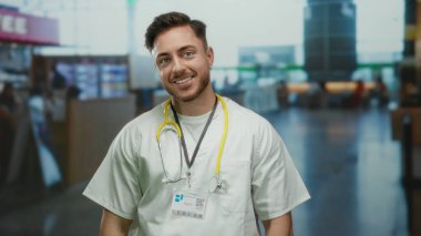 Havaalanı terminalinde steteskopla gülümseyen beyaz erkek doktor yoğun bir seyahat ortamında profesyonel ve ulaşılabilir bir tavır sergiliyor..