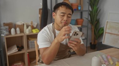 Önlük giyen genç Çinli bir zanaatkar, çanak çömlek atölyesinde eli çenesinde akıllı telefon tutar..