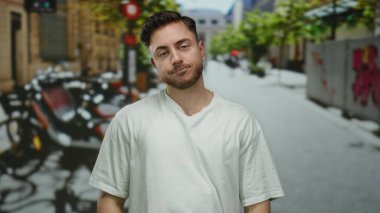 Sakallı genç adam canlı bir şehir caddesinde duruyor, canlı yeşillik ve şehir manzarası ile çevrili, modern açık hava yaşamını yansıtıyor..