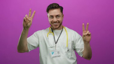 Sakallı genç erkek doktor neşeyle gülümsüyor ve ensesinde steteskopla izole edilmiş pembe arka plana karşı barış işaretleri gösteriyor..