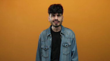 Koyu renk saçlı ve kot ceketli genç adam turuncu bir arkaplanda tuhaf bir ifade sergiliyor..
