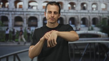 Caddedeki kolezyum binasının önündeki kameraya bakarken bileğine dokunan bir adam, sessiz tefekkür.