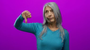 Gri saçlı, mavi tişörtlü yaşlı bir kadın, izole edilmiş pembe duvarlara karşı baş parmak işareti yapıyor..