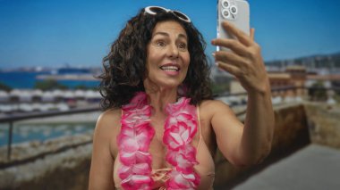 Orta yaşlı, bikinili ve çiçekli bir kadın limanda selfie çekmek için kollarını açmış akıllı telefon tutuyor..