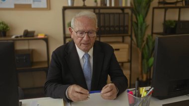 Takım elbiseli ve gözlüklü kıdemli işadamı modern ofis içinde masa başında, elinde bir kalem tutuyordu. Etrafı bitkiler ve ofis malzemeleriyle çevrili, liderlik ve düşünceliliğini temsil ediyordu..