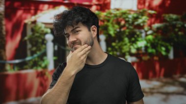 Sakallı genç adam, güneşli bir günde elleri dışarıda saçlarını tarıyor. Kırmızı arka planı bulanık ve bitkiler İspanyol tarzı ve gündelik görünümünü sergiliyor..