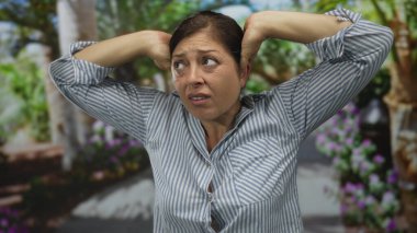 Orta yaşlı İspanyol kadın, güneşli yeşil orman yolunda elleriyle kulaklarını kapatıyor; işitsel rahatsızlık.