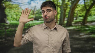 Güneşli ormanda bej gömlekli, gözlüklü, gülümseyen ve kameraya el sallayan bir adam..