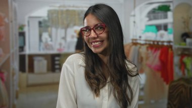 Arka planda renkli kıyafetler olan şık bir mağazada gülümseyen, kırmızı gözlük takan, hareketli ve davetkar bir alışveriş ortamı sergileyen bir kadın..