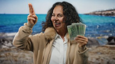 Sahilde tekli banknotlarla gülümseyen bir kadın, zenginliği simgeliyor ve manzaralı okyanus geçmişine karşı jestlerle seyahat ediyor..