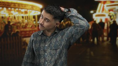 Genç İspanyol adam gece fuarında arka planda atlıkarınca ışıklarıyla eğleniyor, desenli gömlek giyiyor ve canlı park ortamında düşünceli bir şekilde poz veriyor..