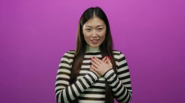 Çizgili gömlek giyen kadın, elleri göğsünde canlı pembe arka plana karşı duran uzun saçlı ve kırmızı tırnaklı çekici ve duygusal bir görünüm sergiliyor..